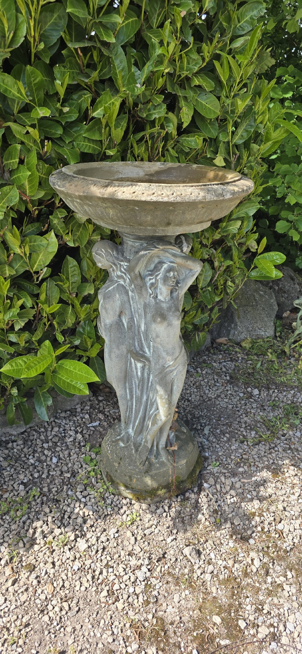 A weathered grey reconstituted stone bird bath featuring a circular top basin resting on a column supported by three draped female figures. View III