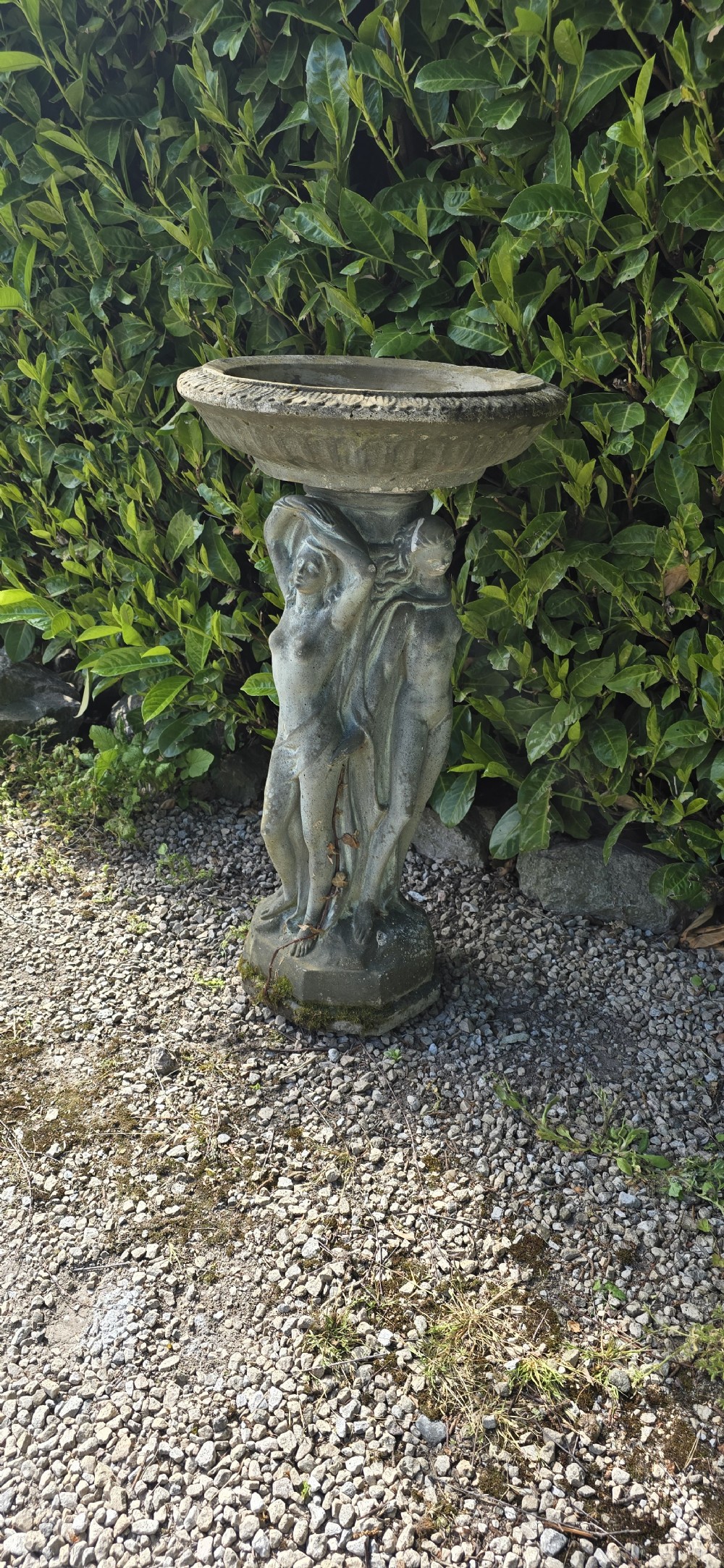 A weathered grey reconstituted stone bird bath featuring a circular top basin resting on a column supported by three draped female figures. View IV