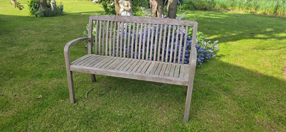 A weathered silver-grey teak garden bench with a slatted back, slatted seat, and curved armrests set upon a lawn. View II