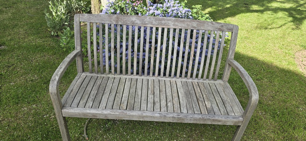 A weathered silver-grey teak garden bench with a slatted back, slatted seat, and curved armrests set upon a lawn. View III