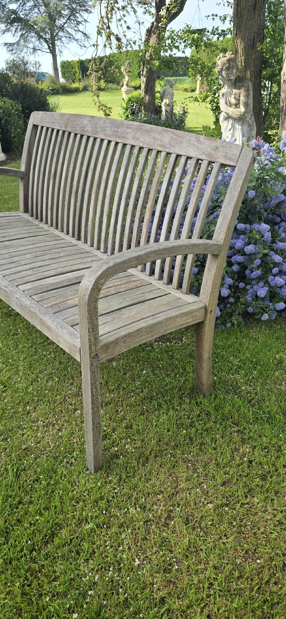 A weathered silver-grey teak garden bench with a slatted back, slatted seat, and curved armrests set upon a lawn. View IV