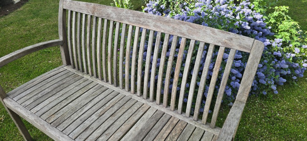 A weathered silver-grey teak garden bench with a slatted back, slatted seat, and curved armrests set upon a lawn. View V