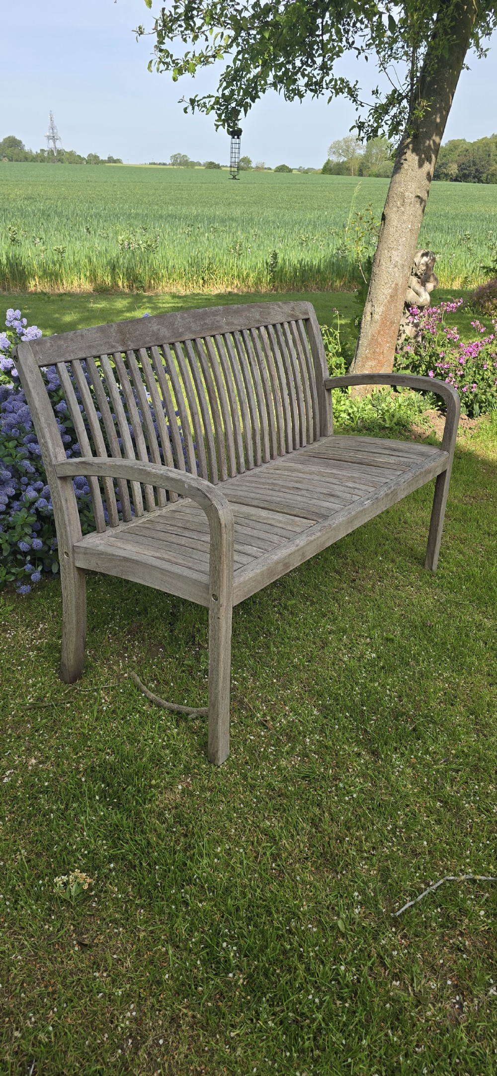 A weathered silver-grey teak garden bench with a slatted back, slatted seat, and curved armrests set upon a lawn. View VI