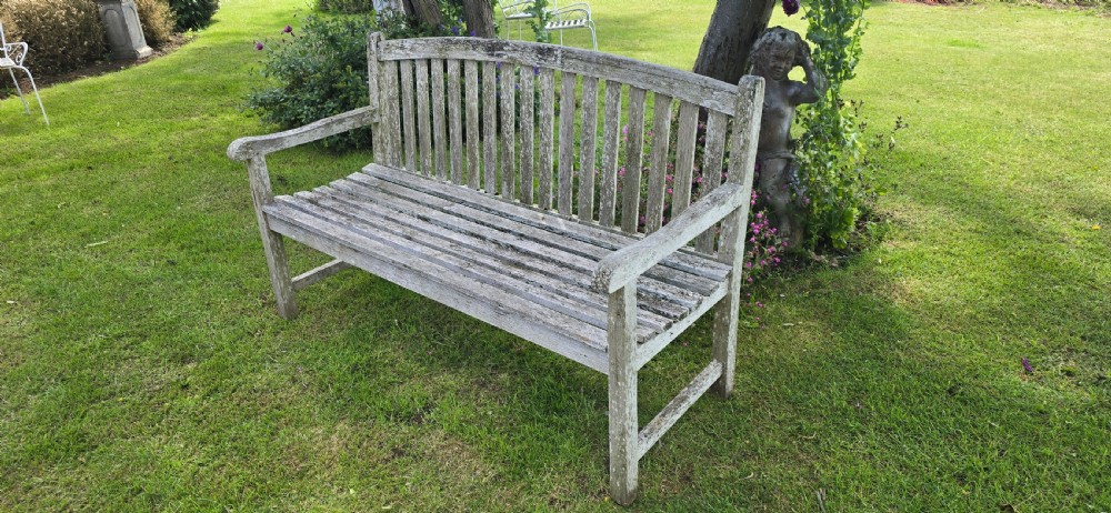 A weathered teak garden bench with a silvered grey patina, featuring vertical back slats, flat armrests, and square legs. View VII