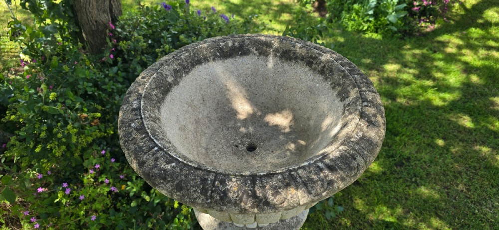 A weathered stone garden urn featuring lobed fluting, elevated on a fluted cylindrical pedestal and square plinth base. View III