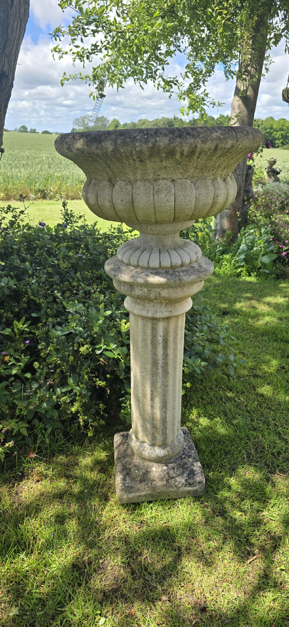 A weathered stone garden urn featuring lobed fluting, elevated on a fluted cylindrical pedestal and square plinth base. View IV