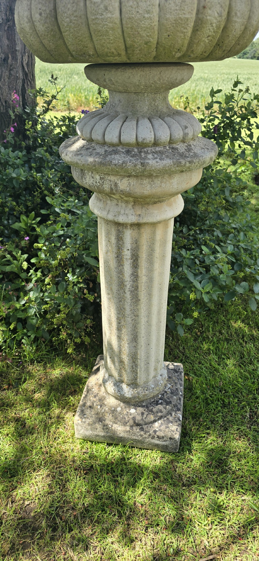A weathered stone garden urn featuring lobed fluting, elevated on a fluted cylindrical pedestal and square plinth base. View V