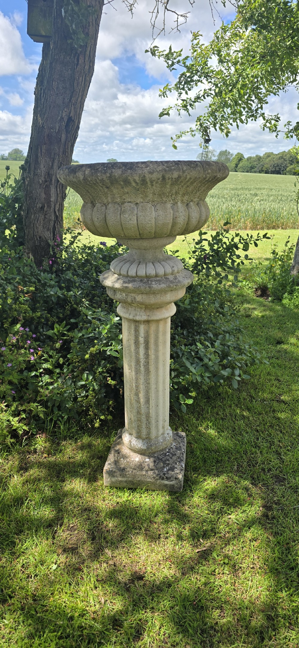 A weathered stone garden urn featuring lobed fluting, elevated on a fluted cylindrical pedestal and square plinth base. View VI
