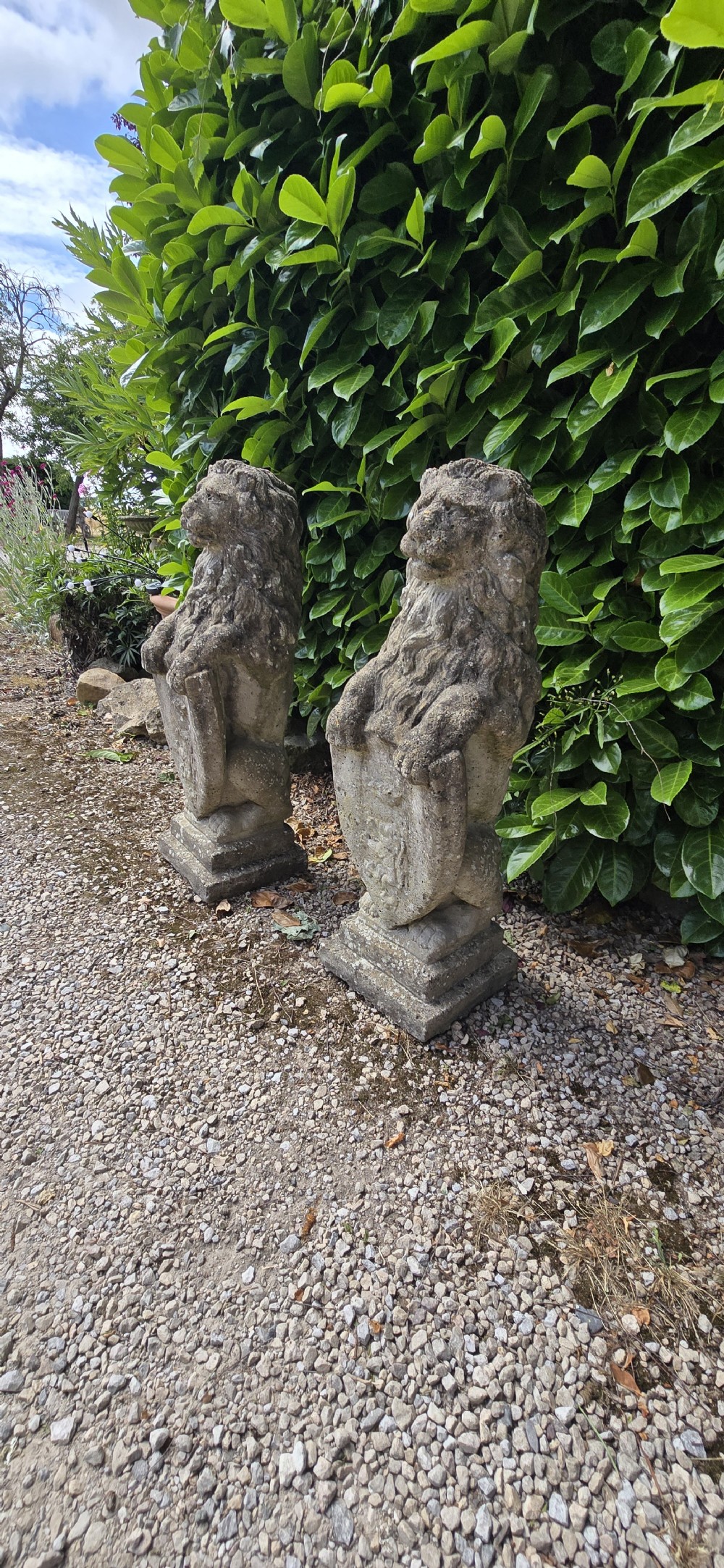 A pair of weathered grey stone garden statues depicting standing lions holding heraldic shields on rectangular bases. View VI
