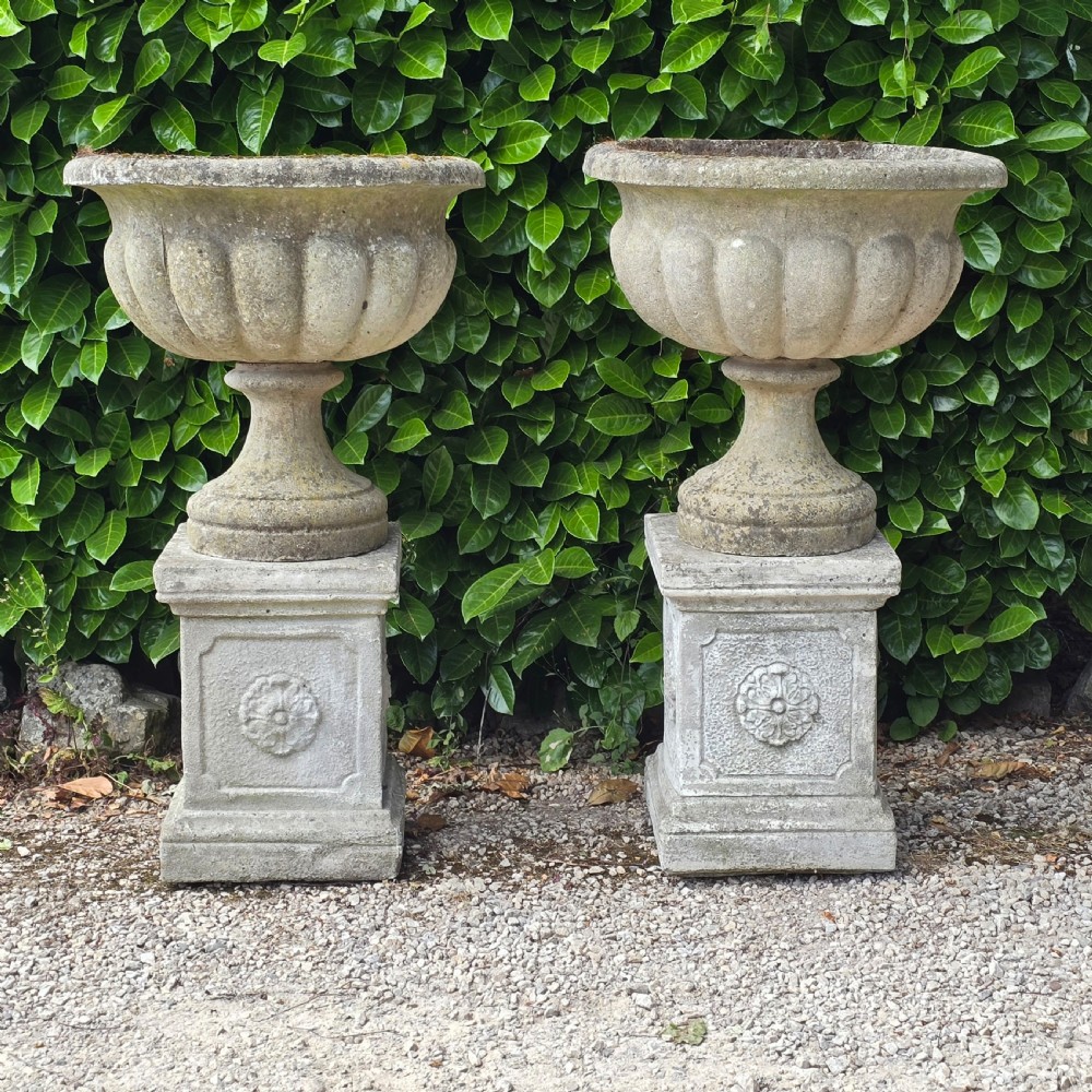 Pair of weathered grey cast stone garden urns featuring fluted bodies and resting on square decorated pedestals.