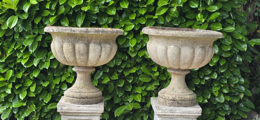 Pair of weathered grey cast stone garden urns featuring fluted bodies and resting on square decorated pedestals. View II