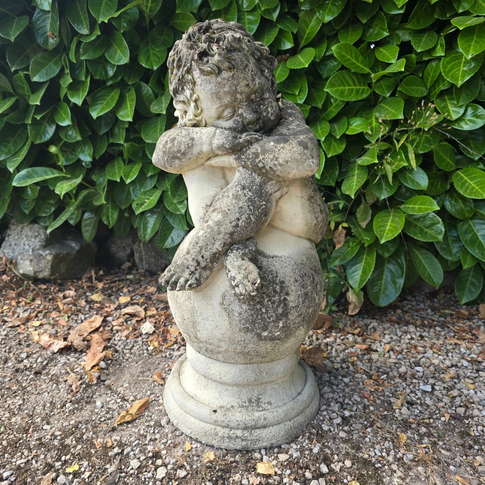 A weathered grey cast stone statue of a sleeping cherub resting its head on crossed arms atop a spherical base and moulded pedestal.