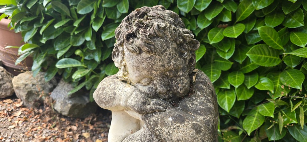 A weathered grey cast stone statue of a sleeping cherub resting its head on crossed arms atop a spherical base and moulded pedestal. View VII