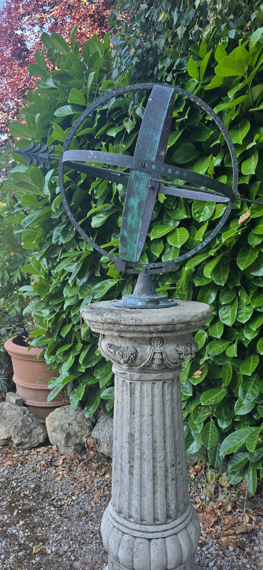 A weathered metal armillary sphere with an arrow gnomon and verdigris patina mounted atop a carved fluted stone pedestal. View VI
