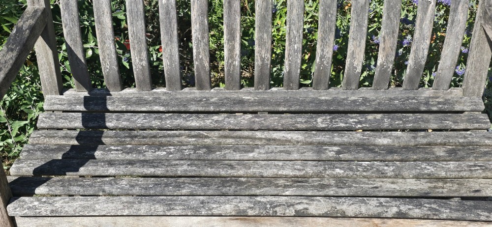 A weathered teak garden bench featuring a vertical slatted backrest, slatted seat, and straight legs connected by stretchers. View IV
