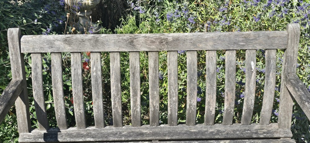 A weathered teak garden bench featuring a vertical slatted backrest, slatted seat, and straight legs connected by stretchers. View V