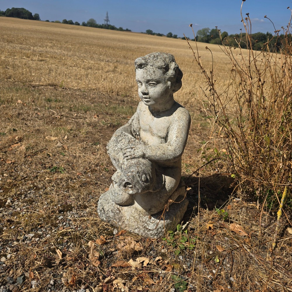 A weathered cast stone garden statue depicting a seated cherub holding a fish.