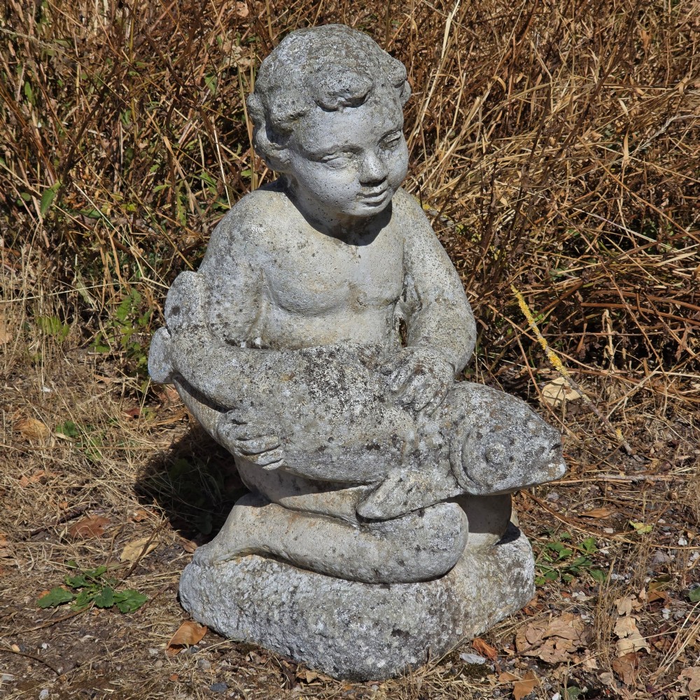 A weathered cast stone garden statue depicting a seated cherub holding a fish. View II