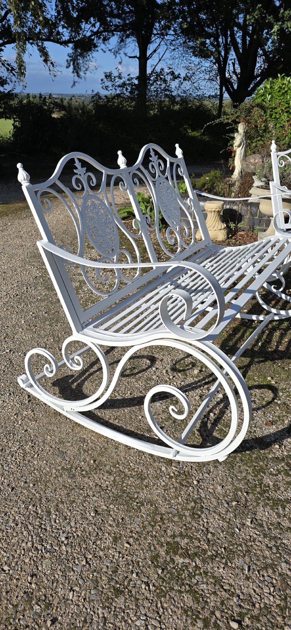 A pair of restored white-painted wrought iron garden rocking benches featuring ornate decorative scrollwork backrests and slatted seats. View V
