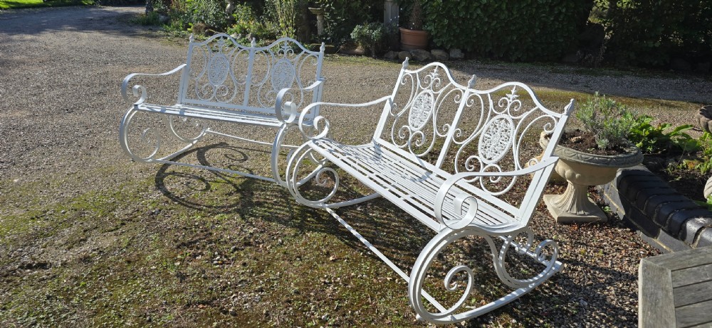 A pair of restored white-painted wrought iron garden rocking benches featuring ornate decorative scrollwork backrests and slatted seats. View X