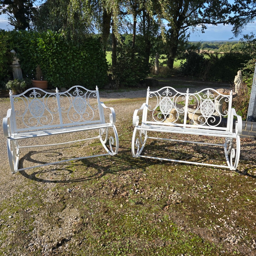 A pair of restored white-painted wrought iron garden rocking benches featuring ornate decorative scrollwork backrests and slatted seats.