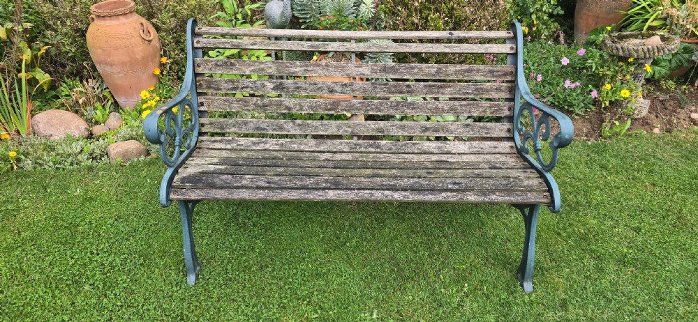 A slatted wooden garden bench with dark green cast iron scrollwork end frames sitting on grass. View III