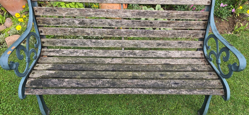 A slatted wooden garden bench with dark green cast iron scrollwork end frames sitting on grass. View IV