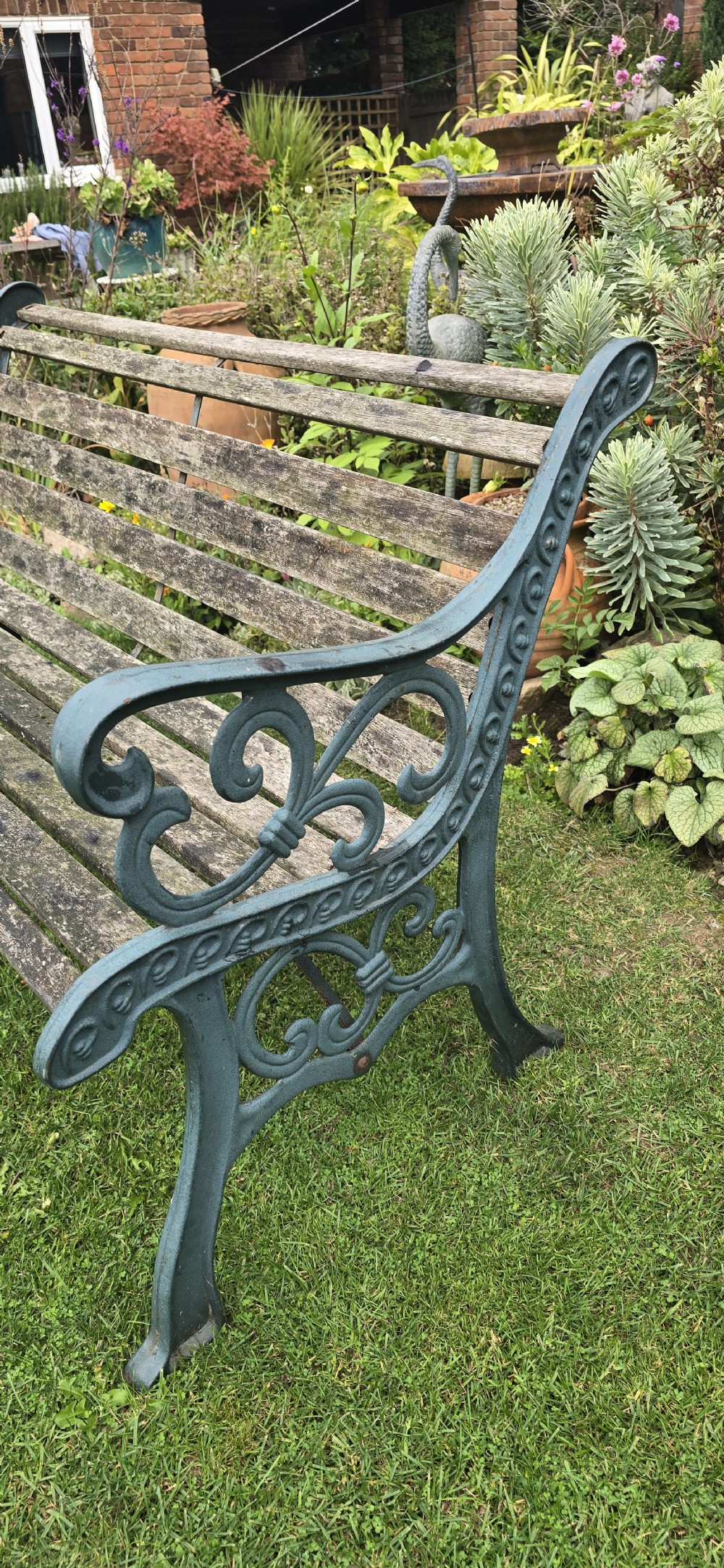 A slatted wooden garden bench with dark green cast iron scrollwork end frames sitting on grass. View VI