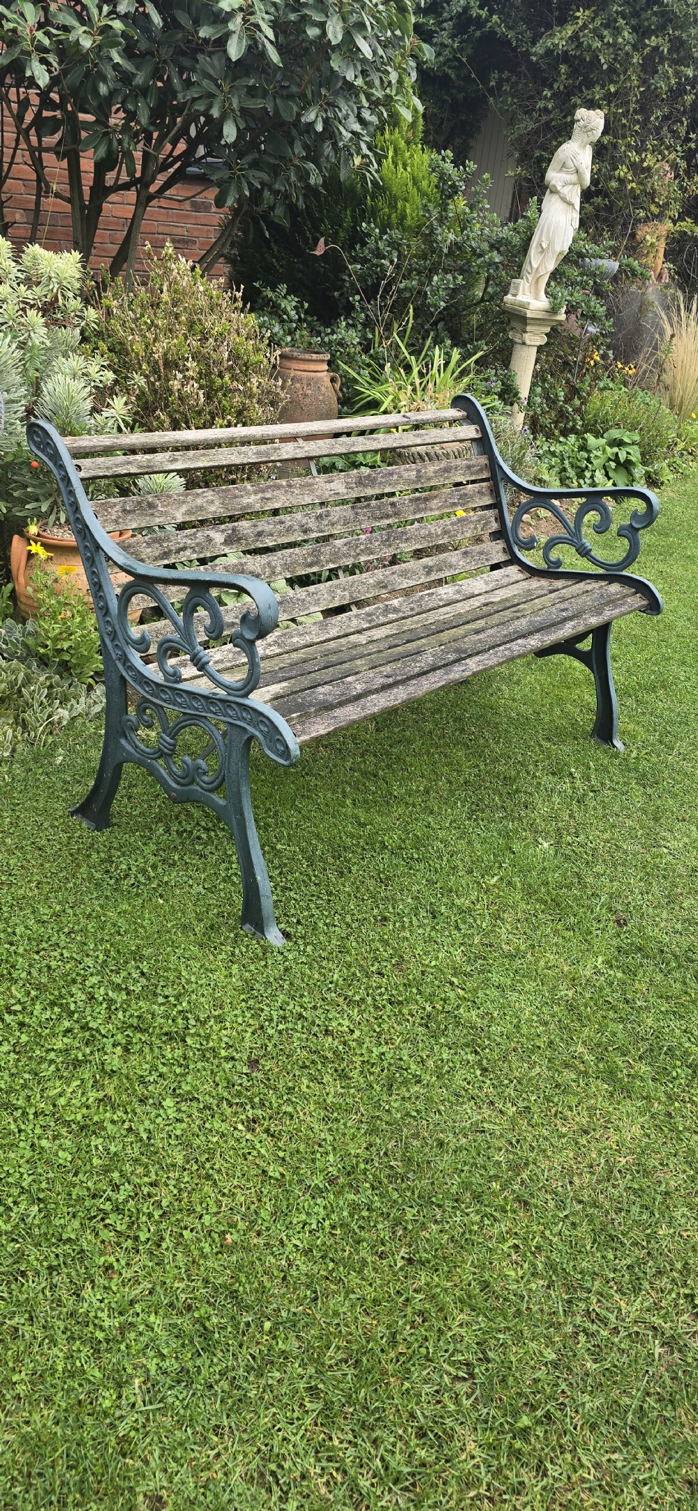 A slatted wooden garden bench with dark green cast iron scrollwork end frames sitting on grass. View VII