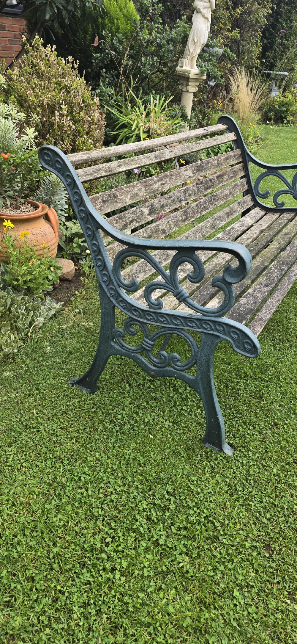 A slatted wooden garden bench with dark green cast iron scrollwork end frames sitting on grass. View VIII