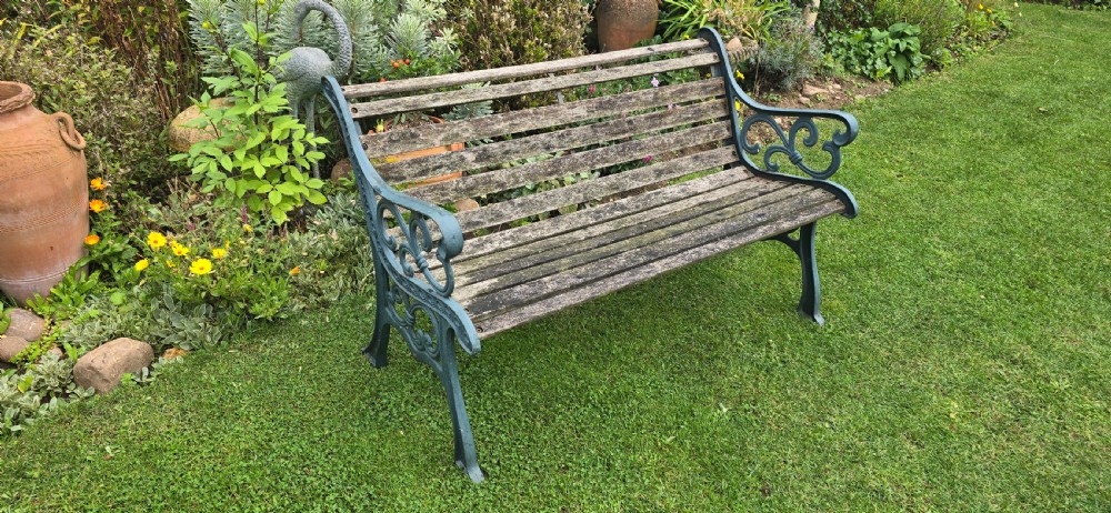 A slatted wooden garden bench with dark green cast iron scrollwork end frames sitting on grass. View IX