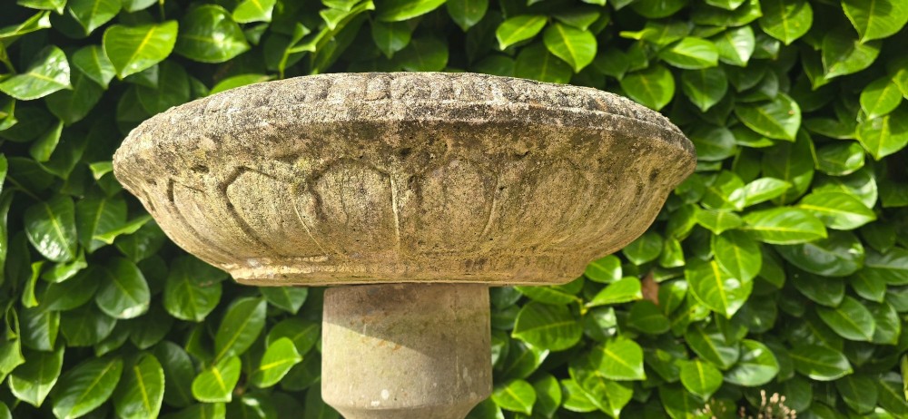 A weathered grey stone bird bath featuring a shallow circular bowl with carved rim detail on a baluster pedestal and round stepped base. View V