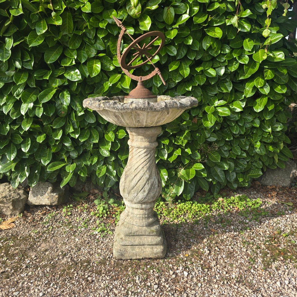 A weathered stone bird bath featuring a spiral-carved pedestal support topped with a rust-brown cast iron armillary sphere.