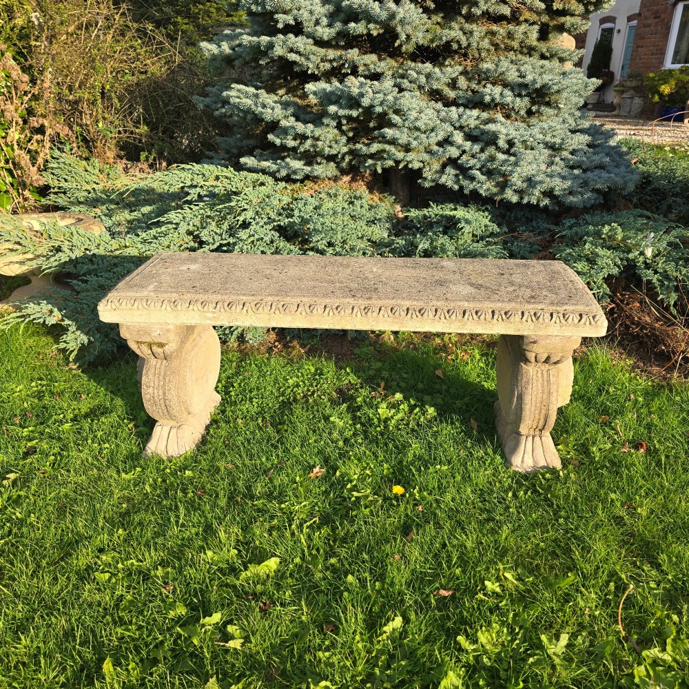 A rectangular weathered stone garden bench featuring carved scroll supports set on lawn grass. View II