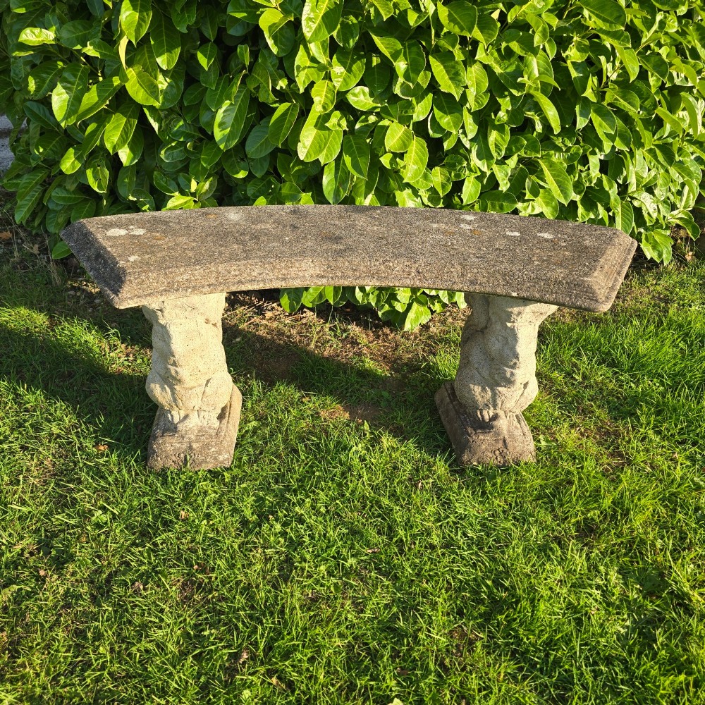 A curved weathered grey stone garden bench featuring a bevelled seat resting on two sculpted animal-form pedestal supports.