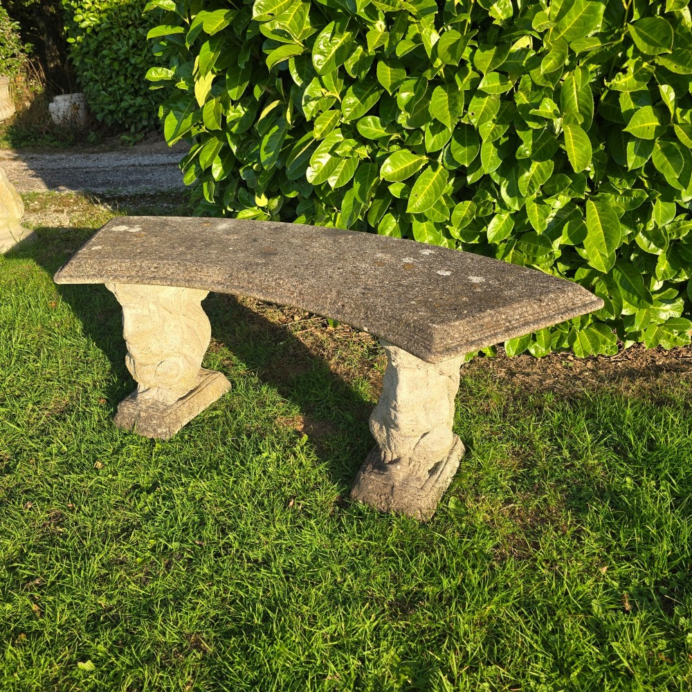 A curved weathered grey stone garden bench featuring a bevelled seat resting on two sculpted animal-form pedestal supports. View IV
