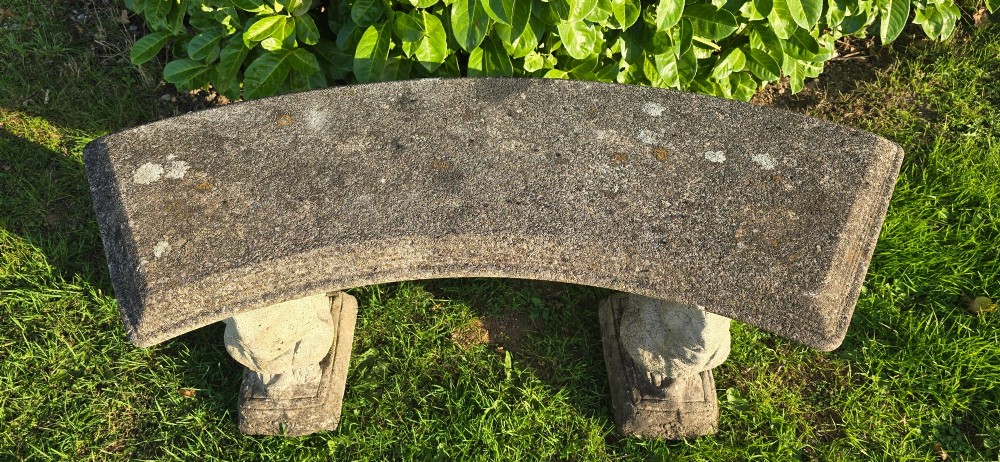 A curved weathered grey stone garden bench featuring a bevelled seat resting on two sculpted animal-form pedestal supports. View VII