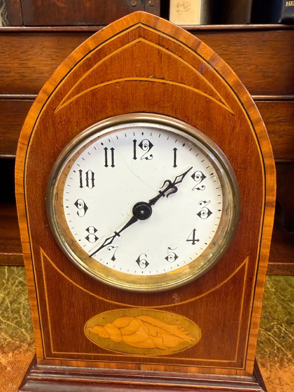 An Edwardian mahogany lancet-top mantel clock featuring satinwood stringing inlay, an oval marquetry shell motif, a white enamel dial with Arabic numerals, and a brass bezel.
