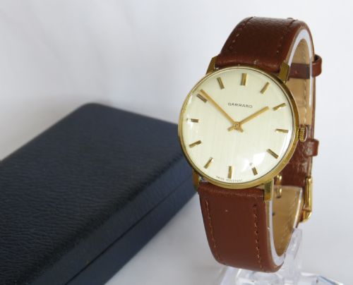 fig 1. A 1960s gents Garrard wristwatch with a gold-toned case, off-white dial, baton markers, and brown leather strap resting inside a navy blue velvet presentation box.