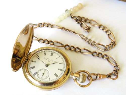 fig 2. A gold-coloured full hunter pocket watch featuring an engraved monogram on the casing, attached to a matching link chain with a decorative toggle.
