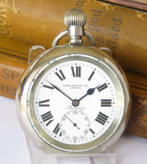 fig 1. An open-face silver-toned pocket watch featuring a white enamel dial with black Roman numerals, subsidiary seconds dial, and text reading John Elkan Ltd London Railway.