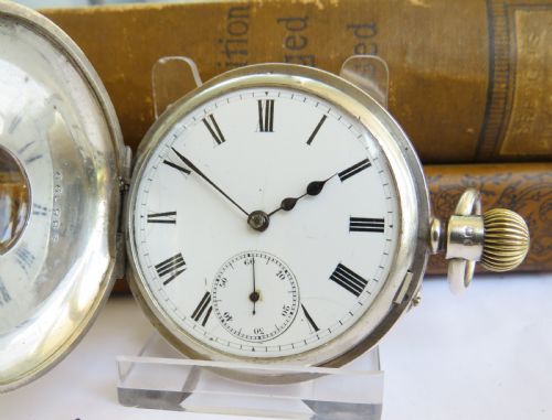 fig 1. An antique sterling silver half hunter pocket watch featuring an engine-turned front case with blue enamel Roman numerals surrounding a central glass window.