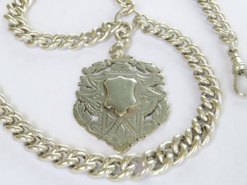 fig 1. An antique silver double Albert pocket watch chain featuring curb links, two swivel dog clips, a central T-bar, and a decorative shield-shaped pendant fob.