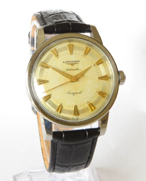 fig 1. A 1957 Longines Conquest automatic wristwatch featuring a stainless steel case, patinated cream dial, gold-tone hour markers, and a black leather strap.