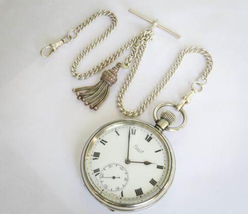 fig 1. An open-face silver-toned pocket watch featuring a white enamel dial, black Roman numerals, a sub-seconds dial, and an attached curb link chain with a tassel fob.
