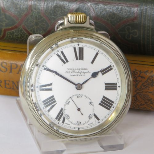 fig 1. An open-face pocket watch featuring a white enamel dial with black Roman numerals, blued steel hands, a subsidiary seconds dial, and a silver-toned metal case with a gold-coloured winding crown.