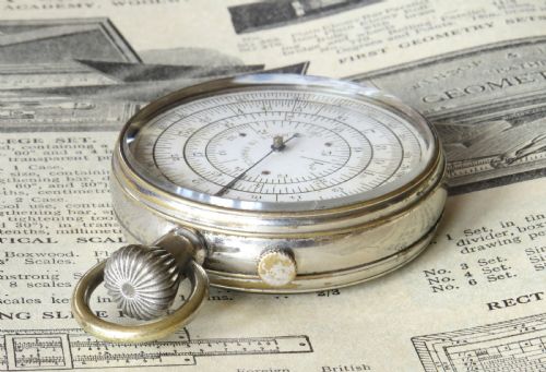 fig 2. A pocket watch styled mechanical circular slide rule with a nickel case, white dial featuring concentric scales, and text reading Calculigraphe H.C. Paris.