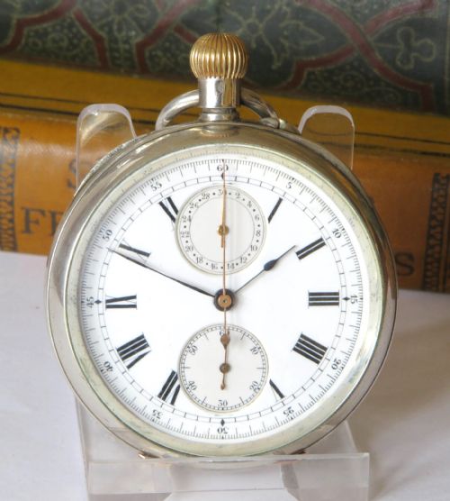 fig 1. An antique silver open-face chronograph pocket watch featuring a white enamel dial with Roman numerals, two sub-dials, and a gold crown.
