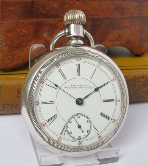 fig 1. An open-face silver pocket watch featuring a white enamel dial with black Roman numerals, red minute tracking, a subsidiary seconds dial, and blued steel hands.