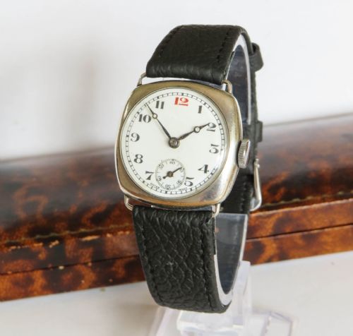 fig 1. An antique 1920s cushion-shaped sterling silver wristwatch with a white enamel dial, black Arabic numerals, a red twelve, and a black leather strap, presented in an open fitted velvet box.